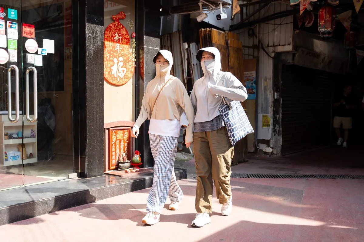 Two women walking down a sunlit street wearing wide-brimmed hats and long-sleeved hooded tops that cover the face and arms